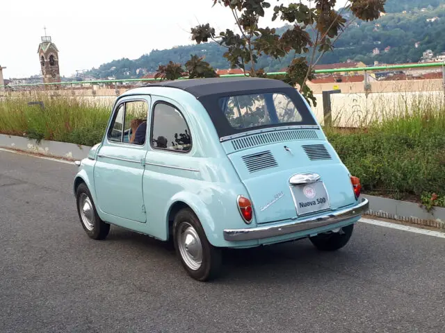 Een lichtblauwe vintage Fiat 500-auto met een zwart dak staat geparkeerd op een weg met groen en een verre klokkentoren op de achtergrond, die de essentie van klassieke auto's vastlegt.