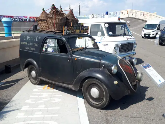 Een oude zwarte auto met manden op het dak staat geparkeerd voor een vintage politiebusje. Op de zijkant staat een tekst in het Italiaans geschreven. Naast de auto staat een uithangbord met waarschijnlijk details over deze klassieke Fiat-modellengespot in de stad.