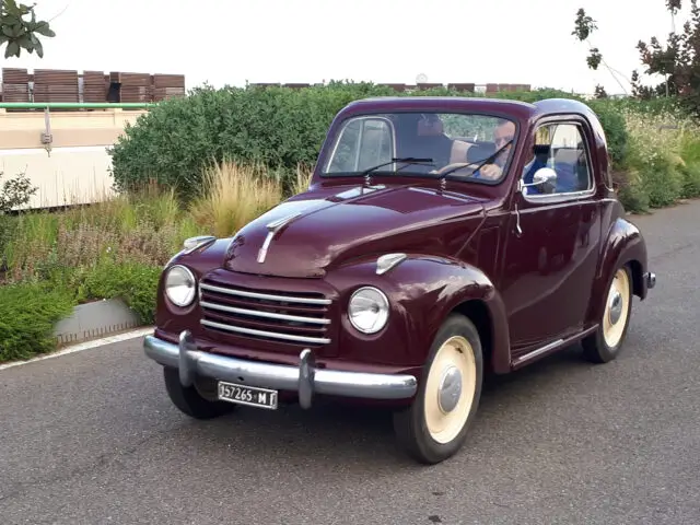 Een kastanjebruine vintage auto met crèmekleurige wielen rijdt op een weg omgeven door groen. Gespot: Een glimp van één van de klassieke Fiat modellen met kenteken "957265 MI".