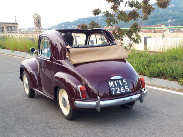 Een vintage kastanjebruine klassieke Fiat met een gedeeltelijk naar beneden gevouwen beige cabriokap en een Italiaans kenteken rijdt door een straat vol groen en verre gebouwen.