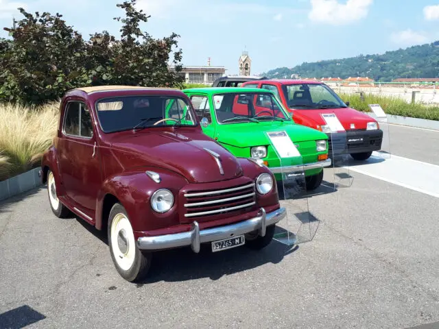 Drie oldtimers, waaronder een klassieke Fiat, een kastanjebruine, een groene en een rode, staan tentoongesteld op een parkeerplaats buiten met informatieborden ernaast. Een schilderachtige achtergrond bestaat uit bomen en een klokkentoren in de verte.