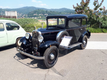 Op een zonnige dag met een bergachtige achtergrond staat een zwarte vintage auto, die doet denken aan klassieke Fiat-modellen, buiten geparkeerd. Links is een gedeeltelijk zicht op een andere auto zichtbaar.