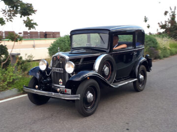 Een zwarte vintage auto, die doet denken aan klassieke Fiat-modellen, met een ronde motorkap en spatborden en een grille met horizontale lijnen, staat gespot op een verharde weg omgeven door weelderig groen.