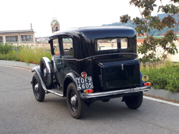 Een vintage zwarte auto met kenteken "TOF 4605" rijdt op een verharde weg, omgeven door groen en gebouwen op de achtergrond. Deze gespot Fiat is een opvallende verschijning onder de klassieke Fiat-modellen en legt de essentie van tijdloos autodesign vast.