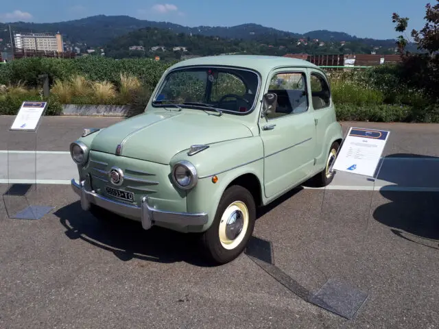 Op een zonnige dag staat buiten een lichtgroene oldtimer tentoongesteld, met informatieborden ernaast. Op de achtergrond zijn groene heuvels en gebouwen. Dit klassieke Fiat-model werd door liefhebbers gespot en voegde charme toe aan het schilderachtige uitzicht.