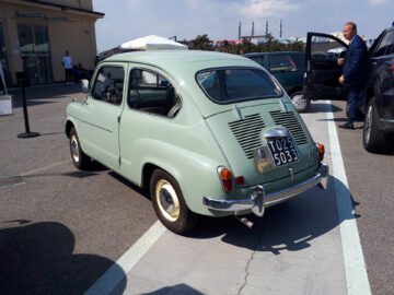 Op een parkeerplaats staat een lichtgroene klassieke Fiat geparkeerd. Een man in pak staat naast een open deur van een nabijgelegen voertuig.