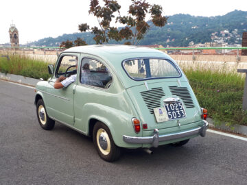 Groene vintage auto, die doet denken aan klassieke Fiat-modellen, met iemands arm tegen het raam, rijdend over een weg met in de verte uitzicht op een stad en heuvels op de achtergrond.