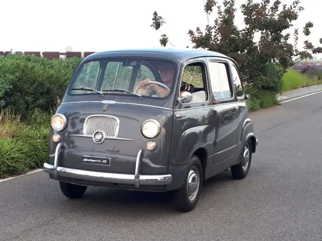 Een vintage grijs busje, dat doet denken aan klassieke Fiat-modellen, rijdt over een weg met groen en struiken op de achtergrond. Het voertuig is voorzien van ronde koplampen en een opvallend embleem op de grille.