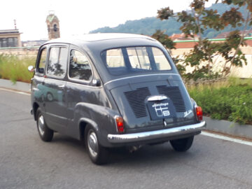 Een klassieke donkergrijze bestelwagen, die doet denken aan klassieke Fiat-modellen, rijdt op een weg met een vage achtergrond van bomen en gebouwen.