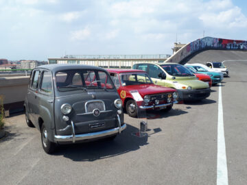 Op een dak met een grijze helling op de achtergrond staat een rij verschillende oldtimers, waaronder enkele klassieke modellen, onder een gedeeltelijk bewolkte hemel.