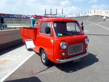 Een rode vintage Fiat-pick-up, onderdeel van de klassieke Fiat-modellen, wordt op een zonnige dag buiten tentoongesteld met informatieve borden ernaast. Gespot op een hellend vlak trekt de vrachtwagen ieders aandacht.