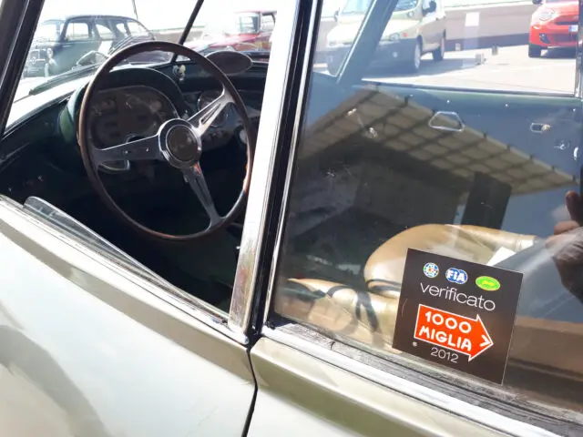 Close-up van het interieur van een klassiek Fiat-model door een raam, met een sticker die deelname aan de Mille Miglia-race van 2012 aangeeft.