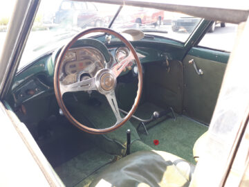 Binnenaanzicht van een klassiek Fiat-model met een houten stuur, analoog dashboard, groene stoelen en versnellingspook. De deur is gesloten en de auto lijkt stil te staan; dit geeft perfect de essentie van vintage elegantie weer. Gespot in onberispelijke staat ondanks de leeftijd.