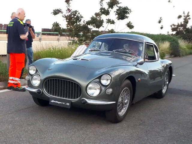 Een vintage sportwagen met een groenachtige tint staat geparkeerd op een weg, met een bordje "Lancia Appia GTE 12" op de grille. Verschillende mensen staan in de buurt en observeren en fotograferen de klassieke Fiat-modellen.