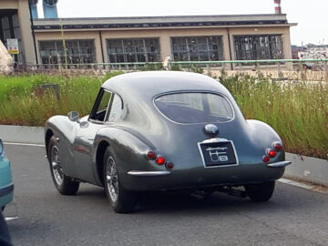 Een vintage grijze klassieke Fiat met een strak, gebogen ontwerp staat geparkeerd langs de weg bij een gebouw. Op het kenteken staat "Heinziger EB." Op de achtergrond zijn struiken en bewegwijzering zichtbaar.