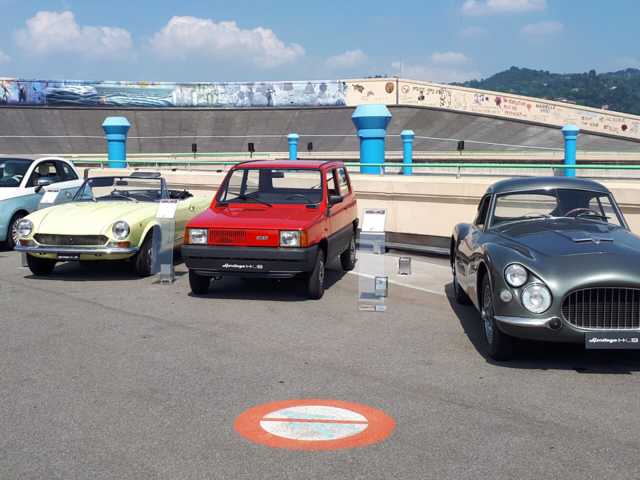 Vier klassieke auto's, waaronder een gele cabriolet, een rode Fiat hatchback en een grijze sportwagen, staan op een zonnige dag buiten tentoongesteld.