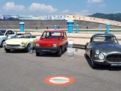 Vier klassieke auto's, waaronder een gele cabriolet, een rode Fiat hatchback en een grijze sportwagen, staan op een zonnige dag buiten tentoongesteld.