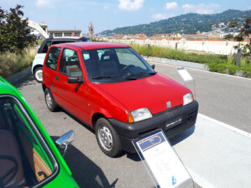 Op een zonnige dag wordt buiten een klassieke Fiat-model tentoongesteld, met een bord en een beschrijving ervoor. Op de achtergrond zijn andere auto's en een stadsgezicht zichtbaar, waardoor een pittoresk stadstafereel ontstaat.