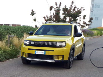 Een geel Fiat-voertuig, een van de klassieke Fiat-modellen, rijdt op een verharde weg met groen en een hoog gebouw op de achtergrond. Rechtsonder op de voorgrond verschijnt de hand van een fietser, alsof hij gevangen zit in een openhartig Gespot-moment.