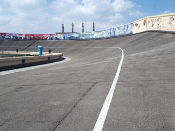 Een lege wielerbaan met schuine bochten en geschilderde muurschilderingen op de muren onder een gedeeltelijk bewolkte hemel, met op de achtergrond een eenzame Fiat geparkeerd.