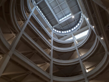 De afbeelding toont een bovenaanzicht van een open atrium met meerdere verdiepingen, ronde balkons en een geometrisch dakraam met rasterpatroon aan de bovenkant. Het natuurlijke licht verlicht de interieurstructuur, wat doet denken aan de architecturale innovatie van Fiat.