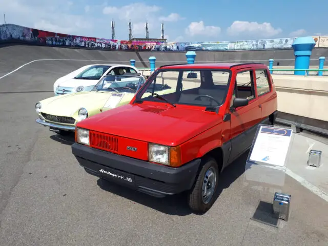 Een felrode hatchback wordt naast een vintage wit klassiek Fiat-model tentoongesteld op een buitencircuit, omringd door informatieborden en een blauwe lucht met wolken op de achtergrond.