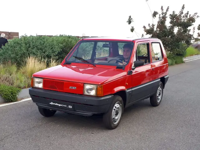 Een rode klassieke Fiat-oldtimer rijdt op een weg met groen op de achtergrond. De auto heeft een hoekige vorm en een klein formaat, wat doet denken aan populaire Fiat-modellen die vaak gespot worden in charmante Europese steden.