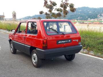 Een rode vintage Fiat Panda 141-serie H.P.6-auto staat geparkeerd op een weg met groen en een stadsgezicht op de achtergrond. Het achteraanzicht benadrukt zijn vierkante vorm en kleine formaat, een echt juweeltje onder de klassieke Fiat-modellen dat op boeiende wijze de charme van klassieke auto's illustreert.