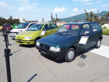 Twee compacte auto's op een parkeerplaats met informatieborden ernaast. De groene auto staat dichter bij de camera, met daarnaast een limoengroene klassieke Fiat. Op de achtergrond is een heuvelachtig landschap zichtbaar.