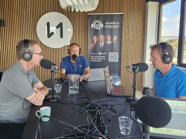 Drie mannen zitten aan een tafel met microfoons, dragen een koptelefoon en zijn bezig met een podcast-opnamesessie. Op de achtergrond is een banner zichtbaar waarin de AutoRAI.nl-podcast wordt gepromoot, waarin automatische concepten in de nieuwste automotive-trends worden besproken.