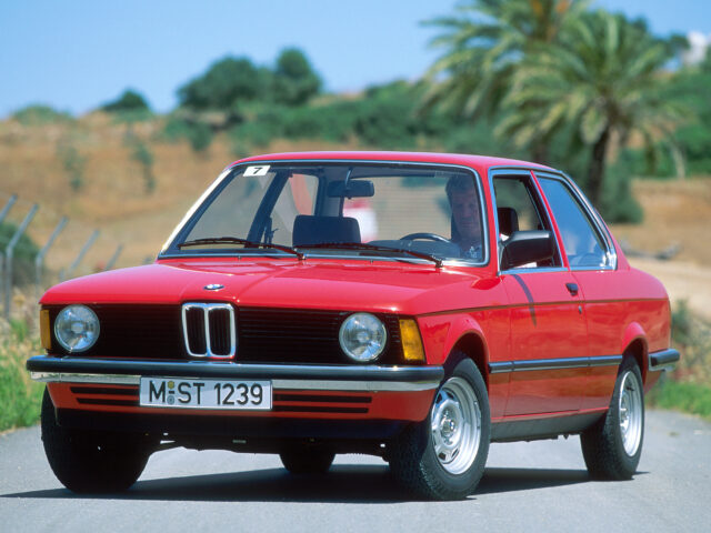 Spotted: A red classic BMW 316 driving down the road, framed by a blurred background of trees and hills on a sunny day. Registration number: M-ST 1239.