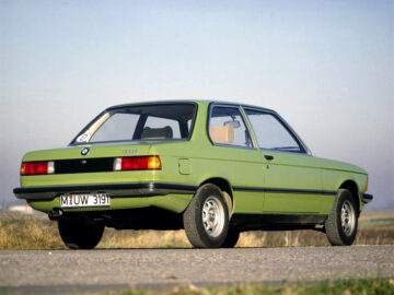 Un BMW 316 antiguo, vestido de verde, está aparcado al borde de una carretera, visto desde la esquina trasera izquierda. Tiene matrícula europea y el horizonte es visible al fondo. La mancha capta el encanto vintage contra un cielo infinito.