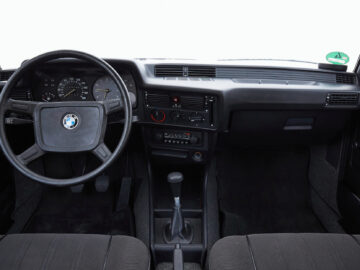 Vista interior de un BMW 316 de época con volante negro con el logotipo de BMW, selector de marchas manual y varios indicadores y botones en el salpicadero. Este antiguo BMW desprende una elegancia atemporal.