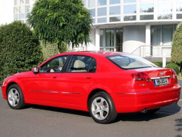 A red Brilliance BS4 sedan is parked on the street in front of a modern building with greenery. The license plate reads 