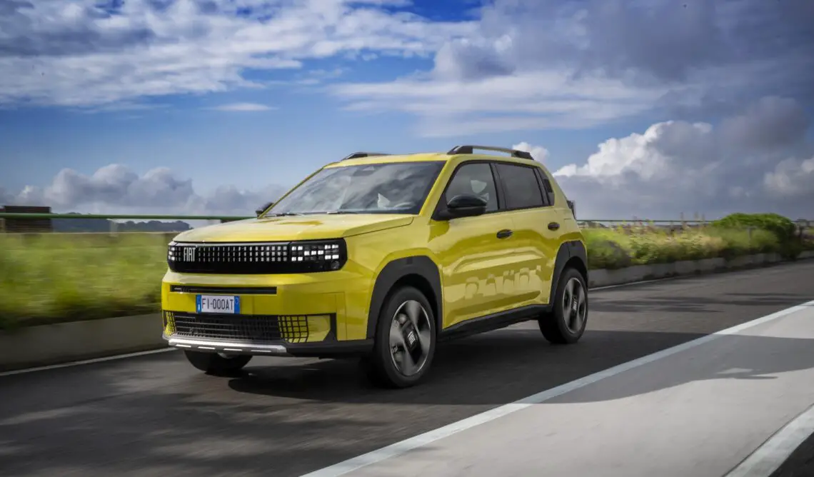 Un SUV Fiat Grande Panda jaune roule sur une route bordée de verdure avec un ciel partiellement nuageux en arrière-plan.