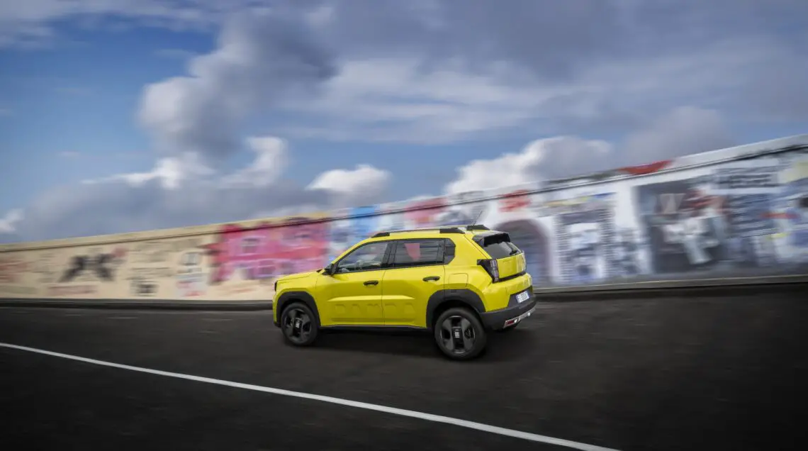 Un SUV Fiat Grande Panda jaune roule sur une route à côté d'un mur couvert de graffitis colorés, sous un ciel partiellement nuageux.