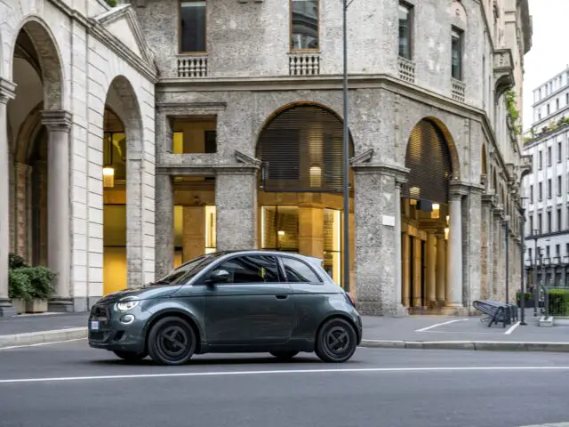 Une petite Fiat Grande Panda vert foncé passe devant un bâtiment historique en pierre avec des arches et de grandes fenêtres dans une rue de la ville.