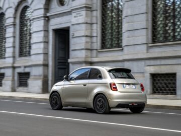 Ein kompakter silberner Fiat Grande Panda fährt eine Straße in der Stadt hinunter, vorbei an einem großen Gebäude mit gewölbten Fenstern und kompliziertem Mauerwerk.