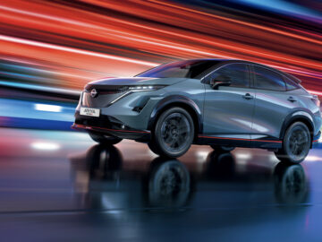 Een gestroomlijnde, moderne Nissan ARIYA SUV in beweging tegen een levendige, dynamische achtergrond met rode en blauwe lichtstrepen. Het metallic grijze voertuig heeft een sportief, aerodynamisch design.