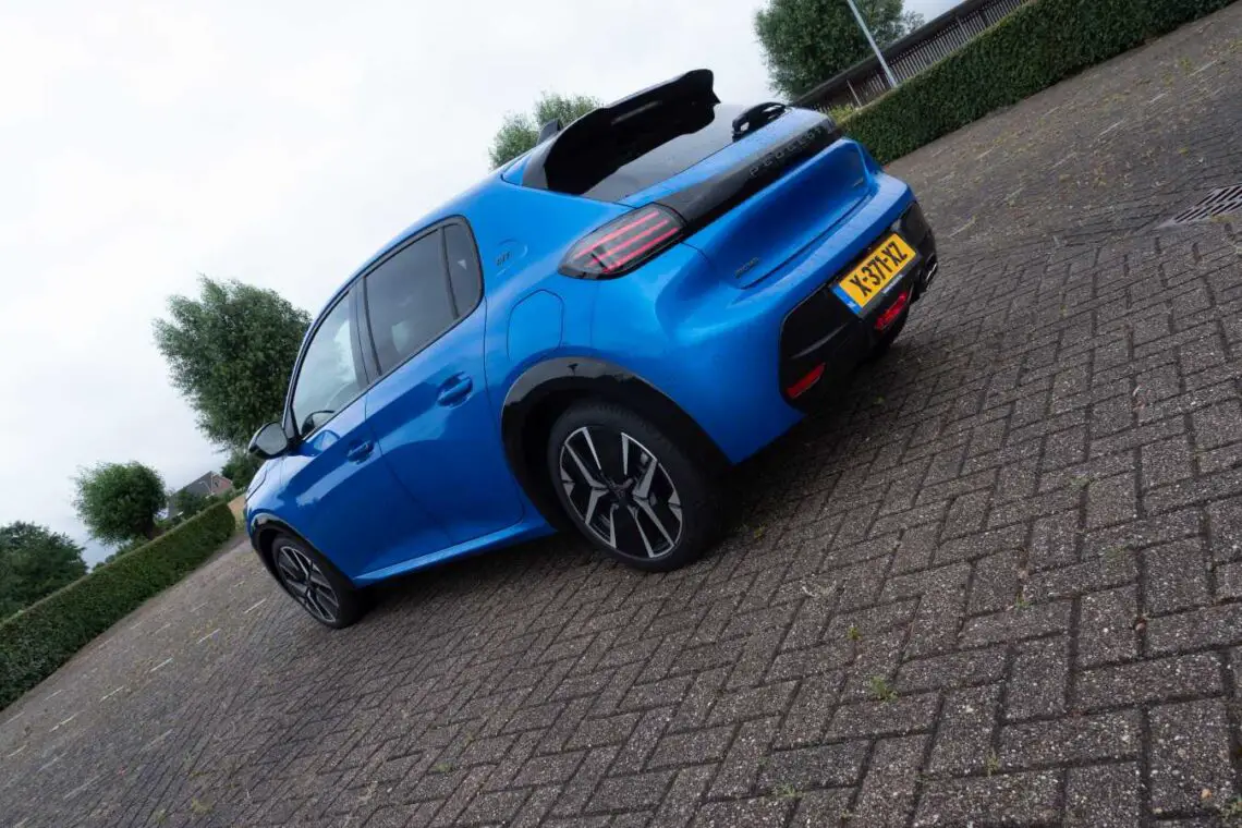 A blue Peugeot 208 Hybrid hatchback is parked in a patterned brick driveway. The car, with the 2024 Dutch license plate and black rims, stands out. Trees and a hedge can be seen in the background.