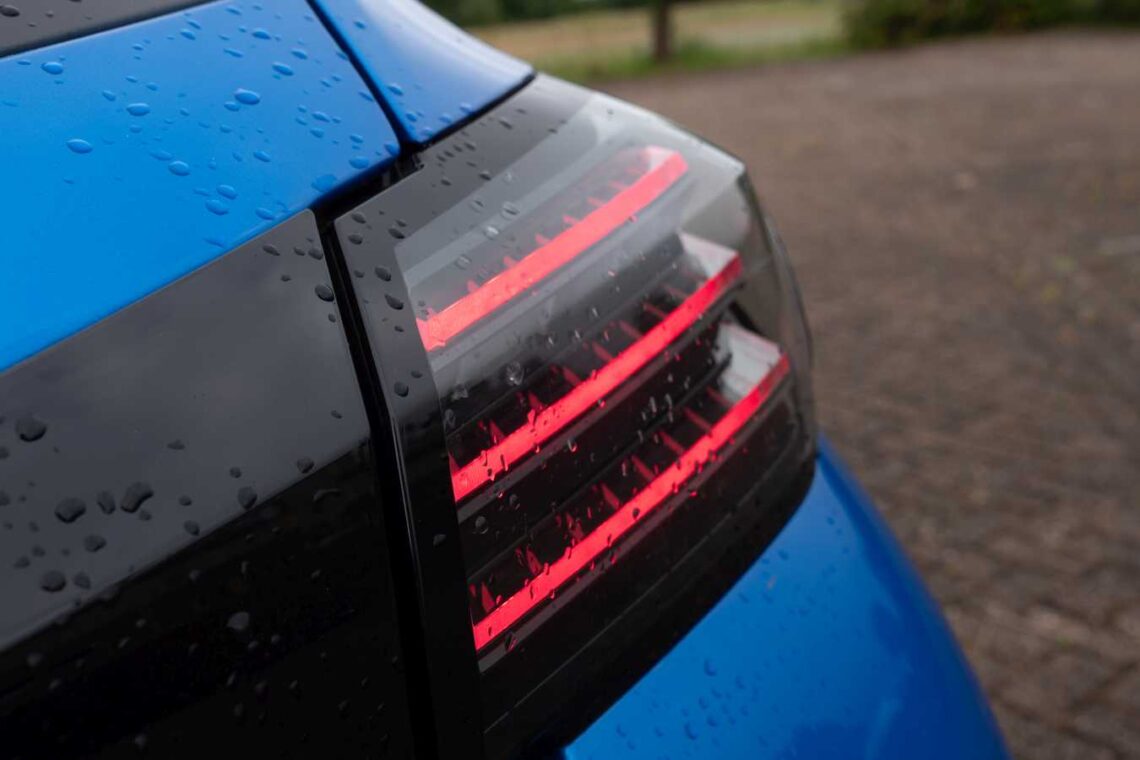 Nahaufnahme des hinteren linken Rücklichts eines blauen Peugeot 208 Hybrid, bedeckt mit Regentropfen, vor einem Hintergrund aus Bürgersteig und Grünflächen, in den Niederlanden.