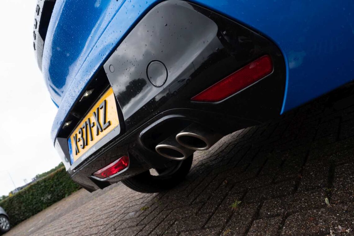 Close-up of the rear of a 2024 Peugeot 208 Hybrid, showing the license plate, dual exhaust pipes and a wet surface, indicating recent rainfall. The car is parked in a brick driveway near a hedge. This model is already popular in the Netherlands.
