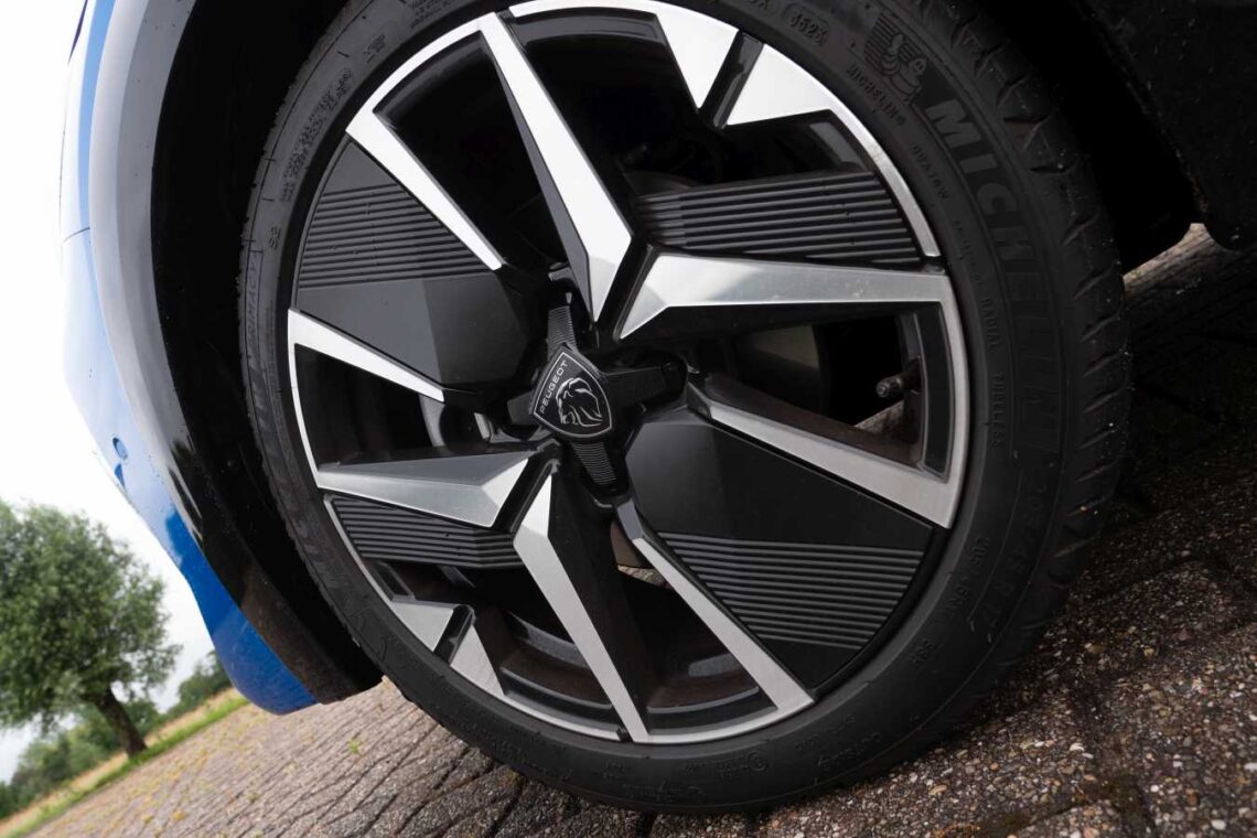A close-up of a car wheel with a black and silver alloy rim with a Peugeot logo in the center. The 2024 Peugeot 208 Hybrid is parked on a paved surface in the Netherlands.