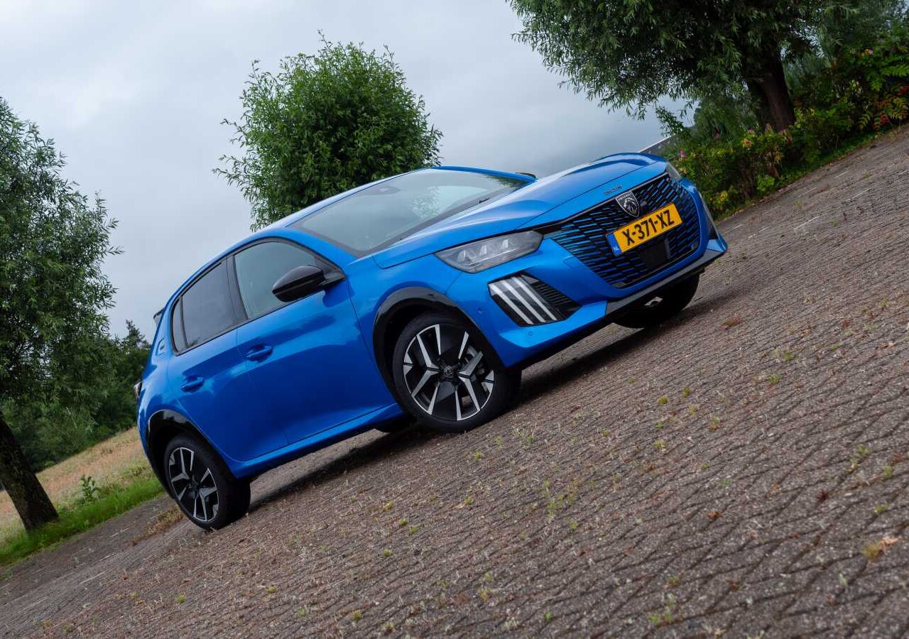Een blauwe Peugeot 208 Hybrid hatchback, populair in Nederland, staat geparkeerd op een verhard oppervlak met bomen op de achtergrond. Op het kenteken van het model uit 2024 staat "K-317-YZL.