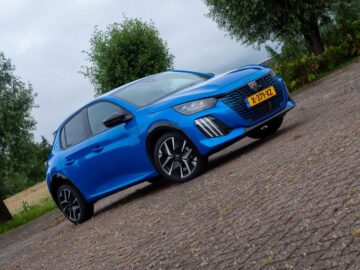 Een blauwe Peugeot 208 Hybrid hatchback, populair in Nederland, staat geparkeerd op een verhard oppervlak met bomen op de achtergrond. Op het kenteken van het model uit 2024 staat "K-317-YZL.