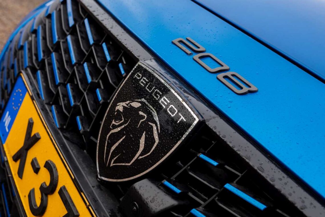Close-up of the grille of a blue Peugeot 208 Hybrid under light rain, showing the iconic Peugeot logo, the 