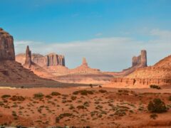 Uitgestrekt woestijnlandschap met rode rotsformaties onder een helderblauwe lucht, met schaarse vegetatie op de voorgrond: een ideale setting voor je volgende woestijnavontuur.