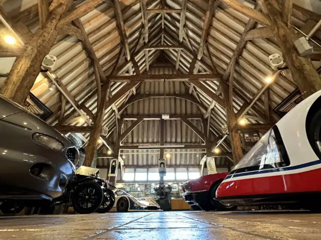 A low-angle view of several Aston Martin cars, displayed in a large, rustic building with wood beams and a high vaulted ceiling. Featuring exposed wooden rafters and natural lighting, the museum's structure embodies the heritage trust of automotive excellence.