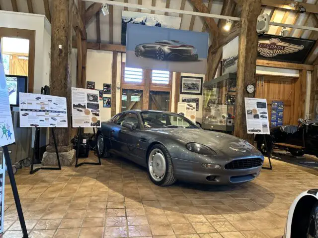 En el interior, se expone un deportivo gris de época con carteles informativos a su alrededor y diversos recuerdos automovilísticos al fondo, creando un efecto sorpresa digno de un reportaje fotográfico del Aston Martin Heritage Trust Museum.