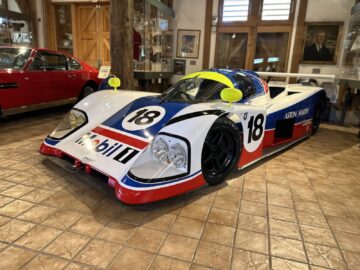 Inside, on a tiled floor is a white race car with blue, red and yellow accents, on which the number 18 is prominently displayed. The car features the Mobil 1 logo and Aston Martin side panels. This striking vehicle is part of the Heritage Trust Museum's latest PHOTOREPORT exhibition.